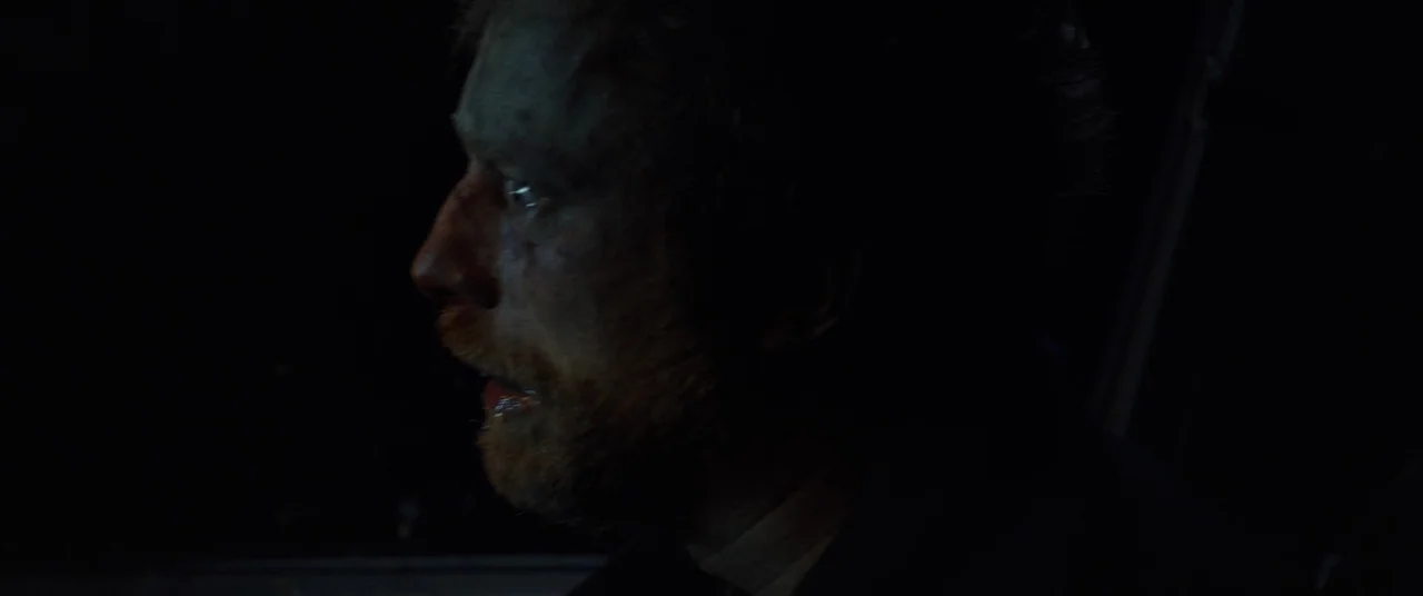 Speak No Evil (2022) movie still: closeup — The interior of a cockpit is dark with minimal ambient light sources. A bearded man with an open mouth…