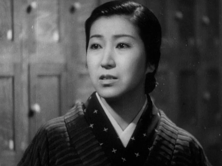 The Most Beautiful (1944) movie still: medium — A woman stands in a dim, wooden architectural interior with traditional Japanese textures. She is…