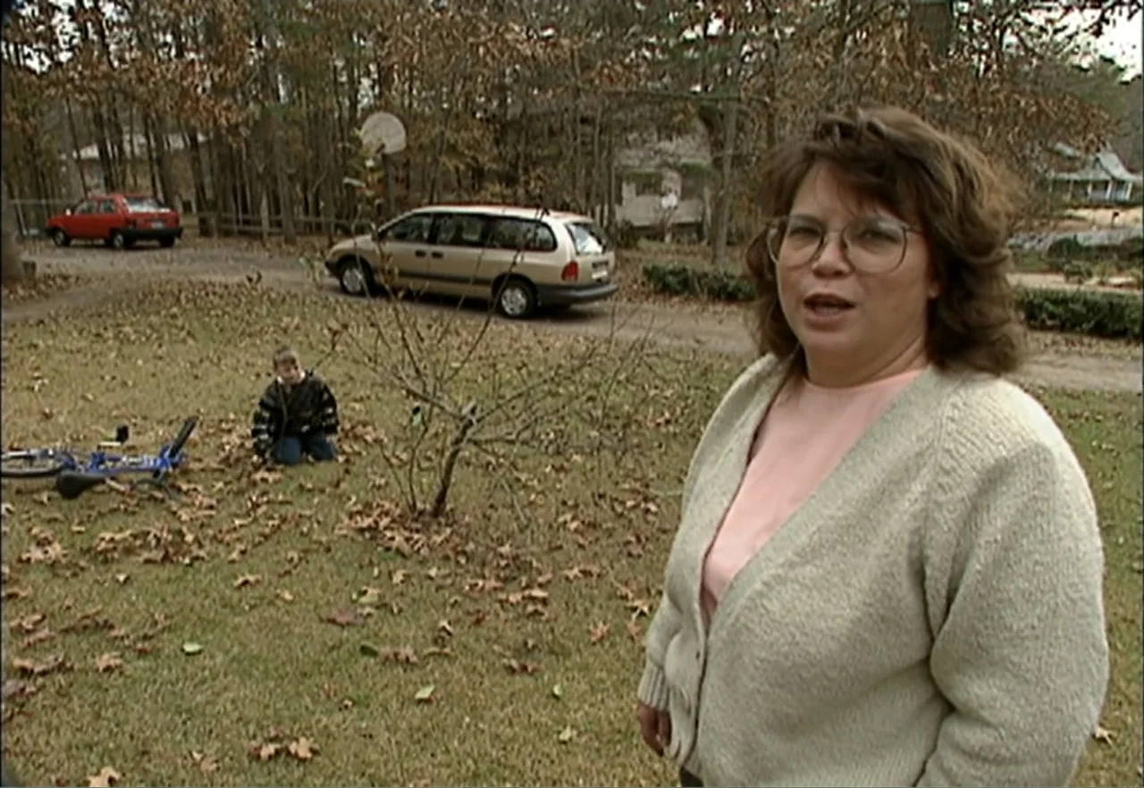 Broken Child (2000) movie still: medium — A residential front yard features a lawn covered with brown autumn leaves and trees. A woman stands in the…