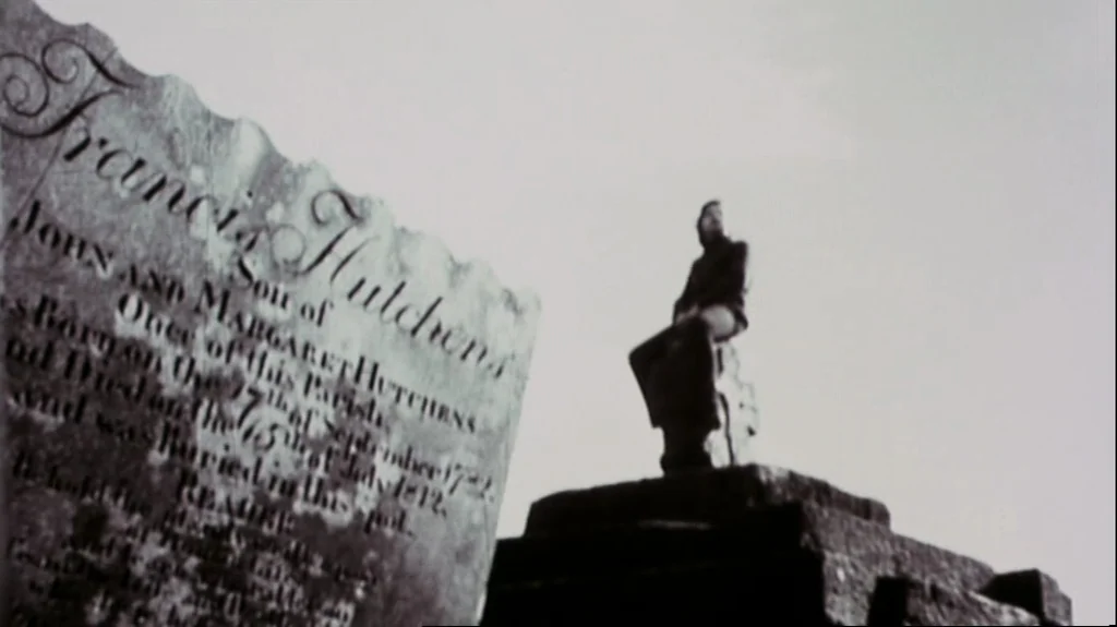 Straw Dogs (1971) movie still: wide — A historic cemetery features weathered stone markers and a raised stone burial vault. A solitary man sits atop…