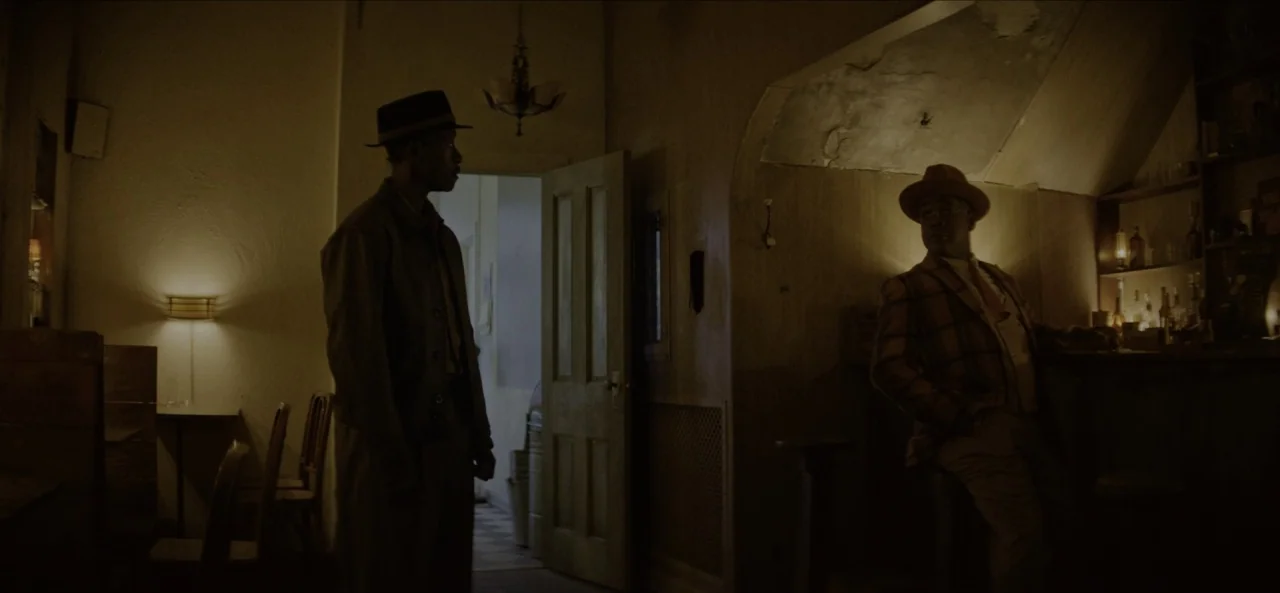 No Sudden Move (2021) movie still: wide — A dimly lit room features wooden walls, a small bar area, and a door frame. Two men in period fedoras…