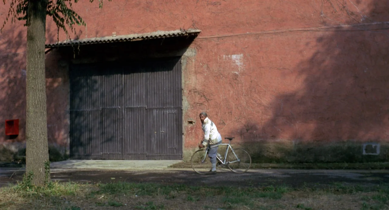 StageFright (1987) movie still: wide — An aged reddish-pink exterior wall serves as a backdrop for a large wooden gate. A man rides a bicycle across…