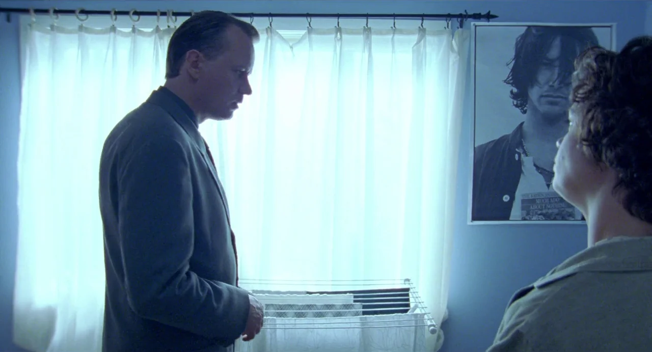 Insomnia (1997) movie still: medium — A room with blue walls features a window with white curtains and a poster. Two men stand facing each other in…