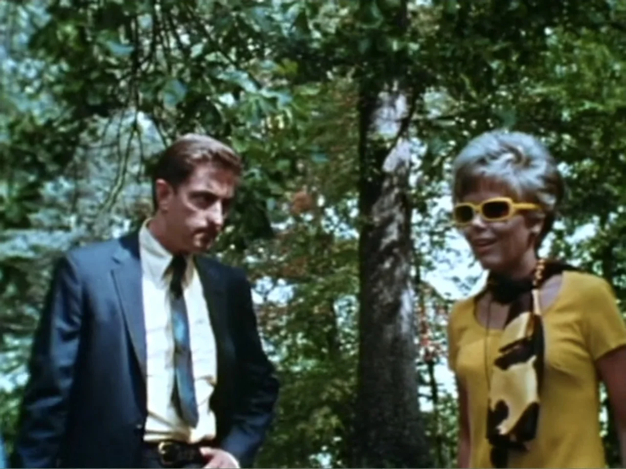 It's Alive (1974) movie still: medium — A dense wooded area with tall trees and dappled light filtering through the leaves. A man in a business suit…