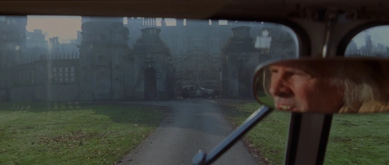 The Haunting (1999) movie still: wide — An imposing stone estate gate sits at the end of a gravel driveway. An older man is partially visible in the…