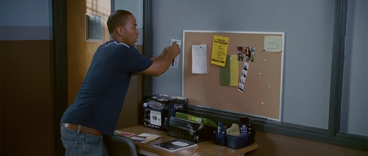 Accepted (2006) movie still: medium — A man stands before a desk in an office featuring a wall-mounted corkboard. He leans forward to pin a small…