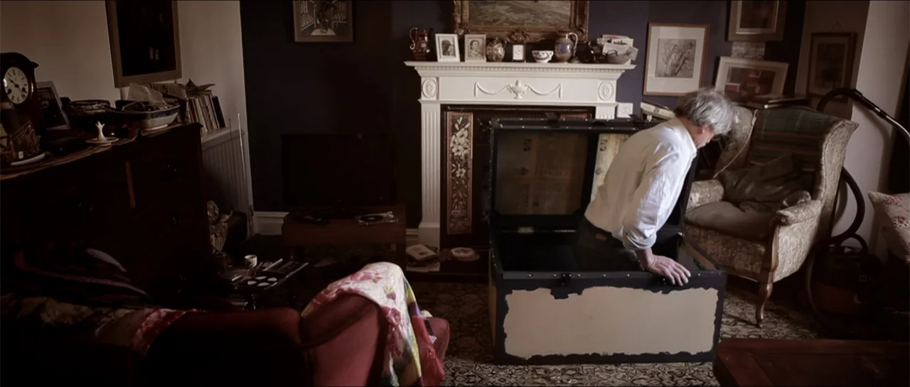 Garnet’s Gold (2014) movie still: medium — A cluttered living room contains a fireplace with mantle decor and multiple framed pictures. An elderly…