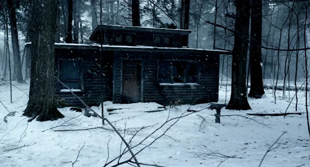 Mama (2013) movie still: wide — A dark wooden cabin sits in a snowy clearing surrounded by tall, leafless trees. Light snow falls gently over the…