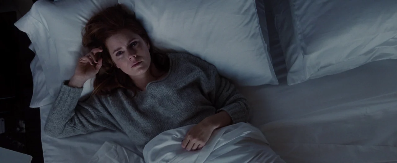 Nocturnal Animals (2016) movie still: medium — A woman lies on white sheets and pillows in a dark, dimly lit environment. She looks directly upward…