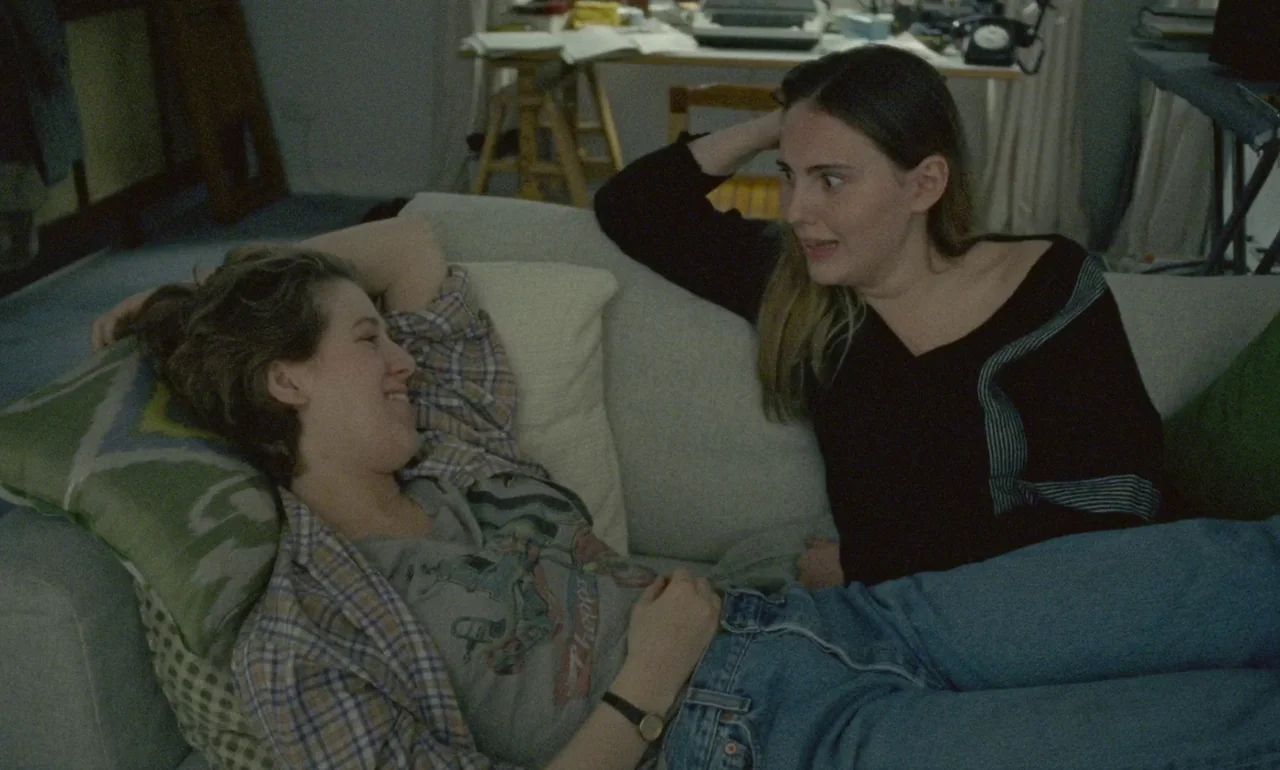 The Souvenir (2019) movie still: medium — A light-colored sofa in a room filled with books and a wooden desk. Two young women lie on the couch while…