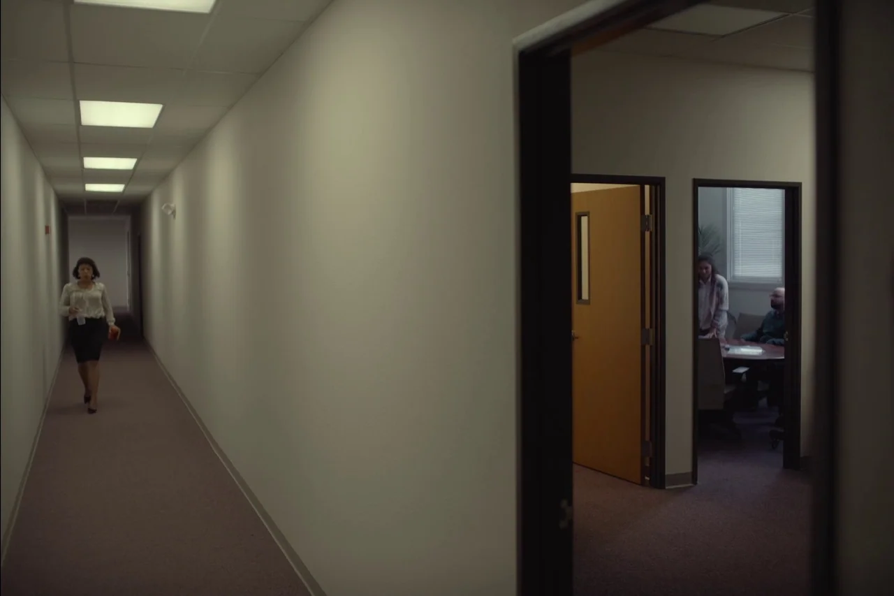 Sometimes I Think About Dying (2023) movie still: wide — A long, sparsely decorated office corridor features plain white walls and grey carpeting. A…