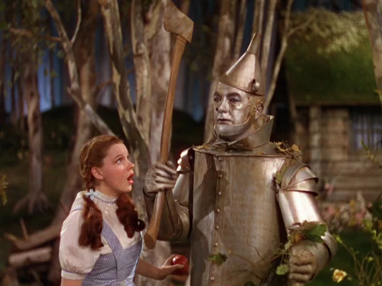 The Wizard Of Oz (1939) movie still: medium — A painted woodland set features artificial trees and a small rustic structure behind. A girl in a…