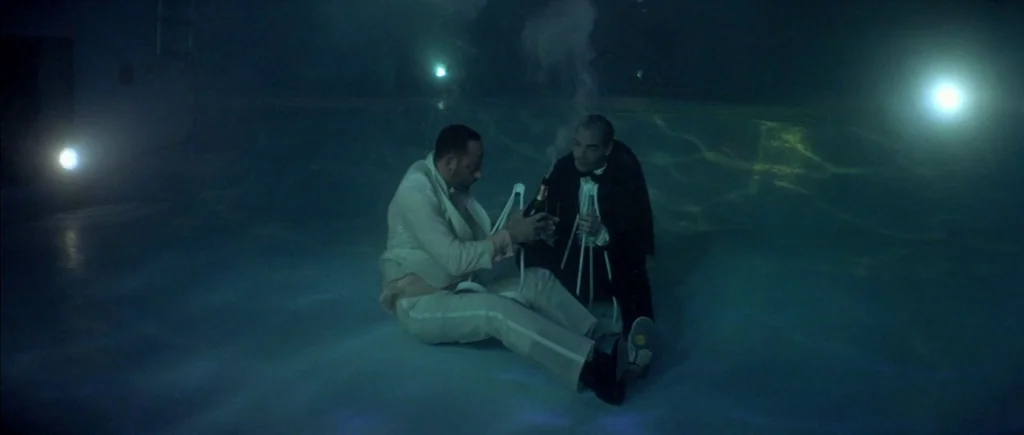 The Big Blue (1988) movie still: wide — Two men in formal tuxedos sit on the floor of a flooded, dark pool. They hold a bottle of champagne and a…