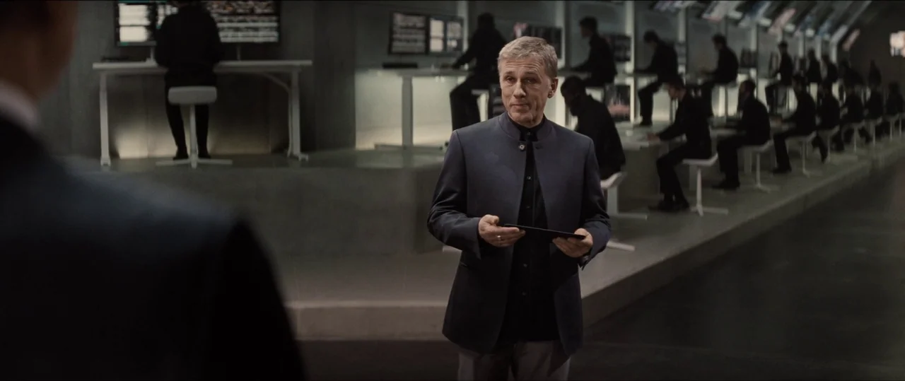 Spectre (2015) movie still: medium — A spacious, modern, dimly lit technology workspace with rows of computer workstations. A man in a dark blazer…