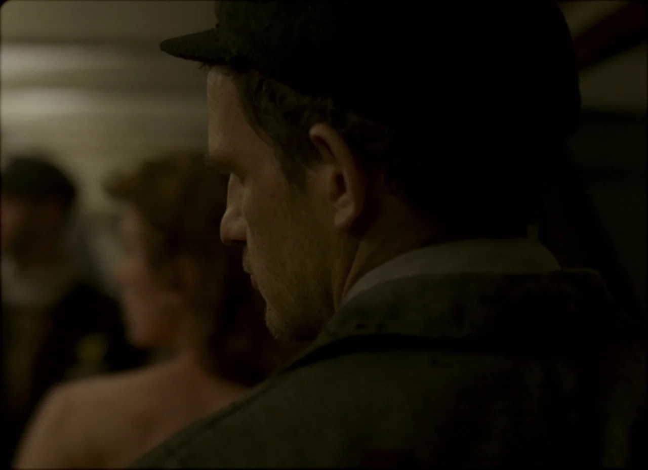 Son of Saul (2015) movie still: closeup — A dark indoor space with blurred figures in the background. A man in a cap is positioned in the foreground.