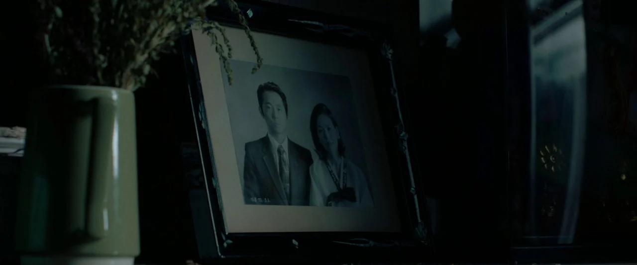 Minari (2020) movie still: closeup — A dark, dimly lit space features a framed black and white portrait of a couple. A green ceramic vessel…