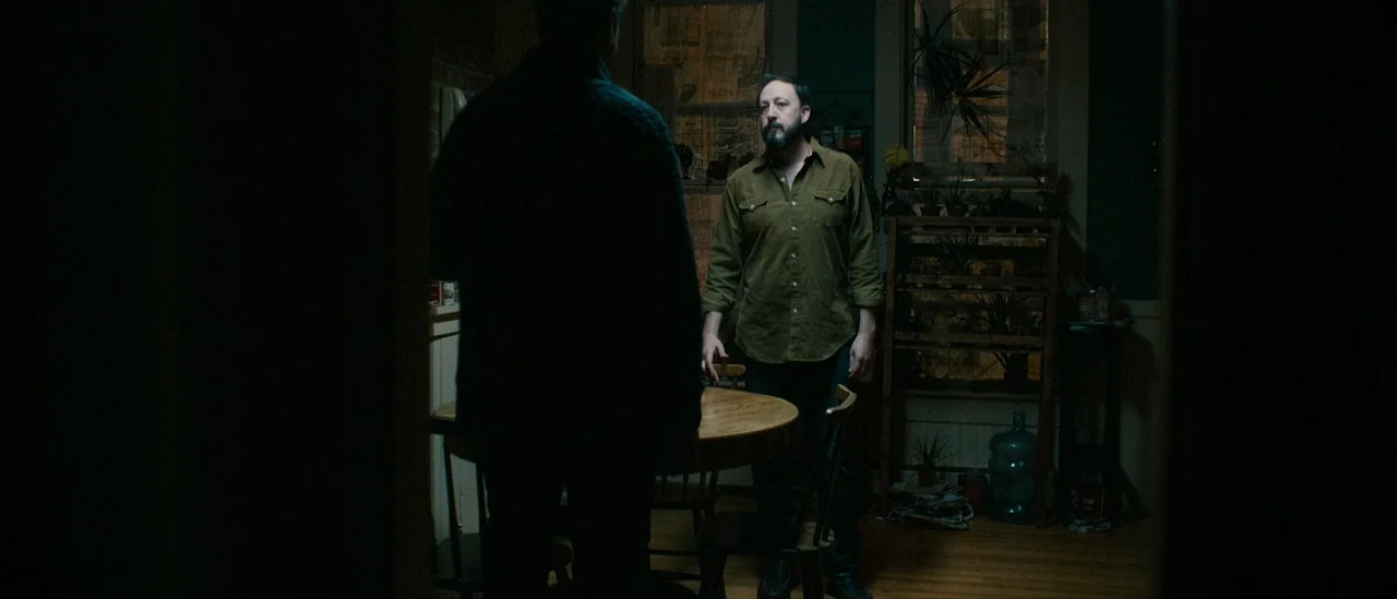 I Trapped The Devil (2019) movie still: medium — A dimly lit room contains a small round wooden table and a plant stand. A man stands near the table…