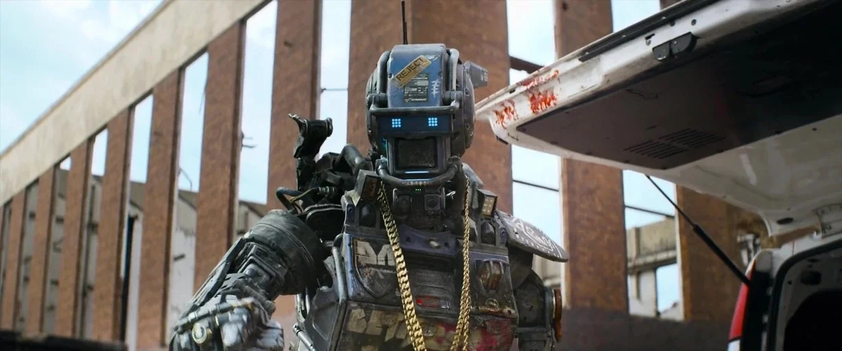 Chappie (2015) movie still: medium — A damaged robot stands before an open vehicle trunk in an unfinished industrial structure. The background…