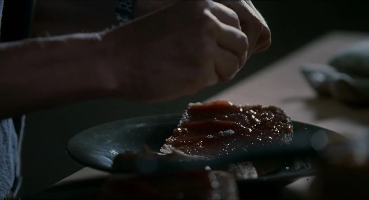 The Ring Two (2005) movie still: closeup — A dark kitchen environment features a wooden table with a plate of toast. A pair of hands spreads a dark…