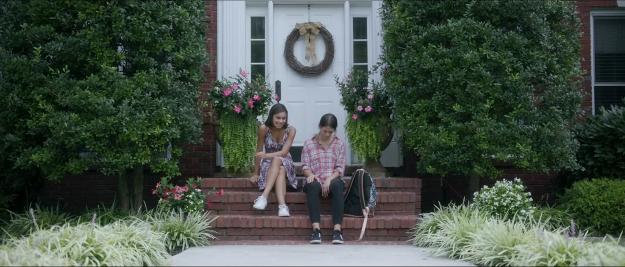 If You're Gone (2019) movie still: fullbody — Two girls sit on the red brick entrance steps of a suburban residential home. Large green bushes and…