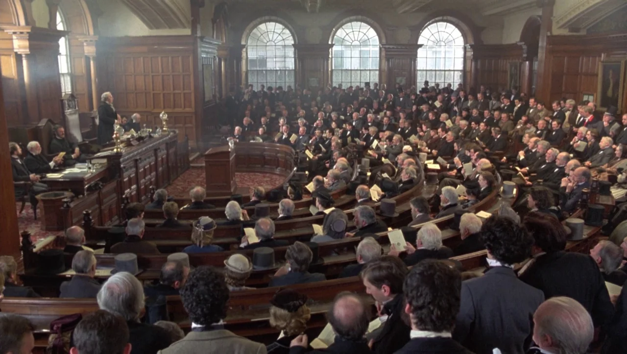 Mountains of the Moon (1990) movie still: wide — A large nineteenth-century legislative hall with tiered wooden seating and arched stained windows.…