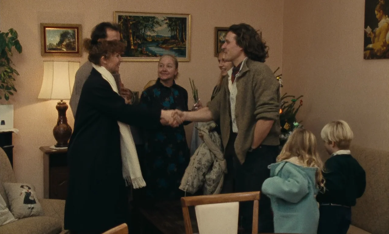 A Tale of Winter (1992) movie still: medium — A domestic living room with tan walls features framed paintings and a table lamp. A group of adults…