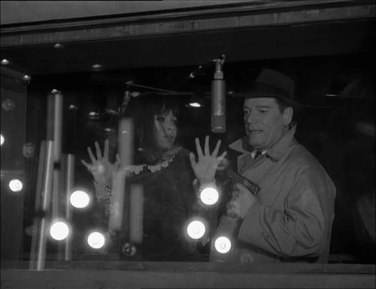Alphaville (1965) movie still: medium — Two individuals occupy a dimly lit space separated from the camera by glass panels. A woman places her hands…