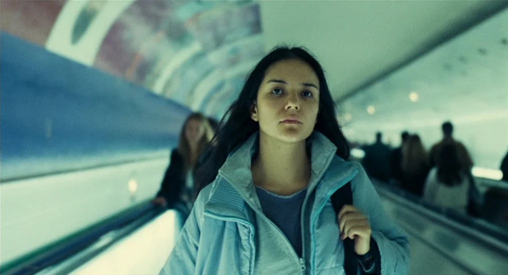 Paris, Je T’Aime (2006) movie still: medium — An interior moving walkway features a long, curved ceiling with light panels. A woman with long dark…