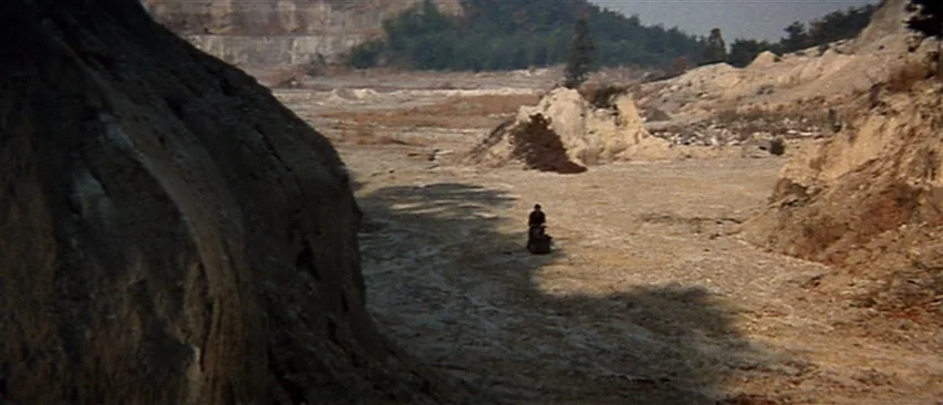 Lone Wolf and Cub: Baby Cart in Peril (1972) movie still: wide — A vast, barren quarry landscape features steep dirt banks and a gravel ground…