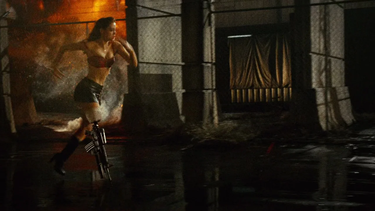 Planet Terror (2007) movie still: fullbody — A dark industrial exterior lot features concrete barriers and orange flames in the distance. A woman…