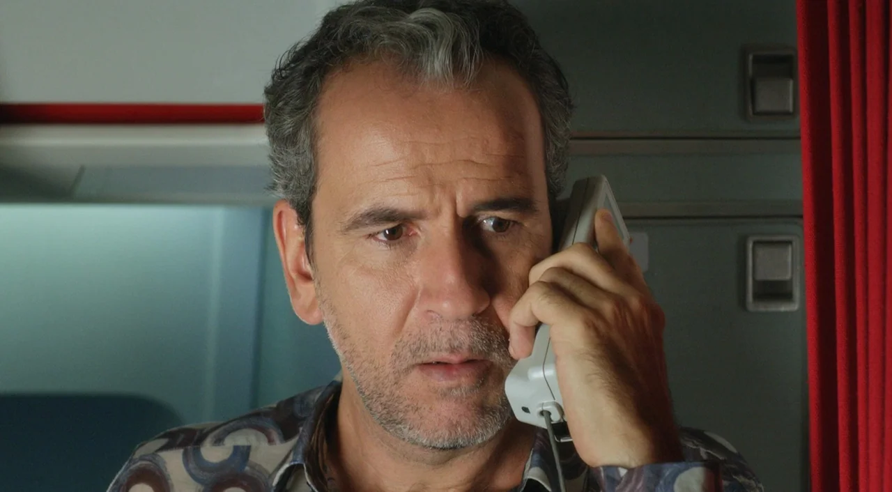 I’m So Excited! (2013) movie still: closeup — An airplane galley features grey metallic wall panels and red curtains on the side. A middle-aged man…