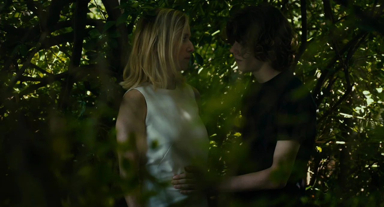 Last Summer (2023) movie still: medium — A dense thicket of green foliage surrounds two people standing in partial shade. The woman in the white top…