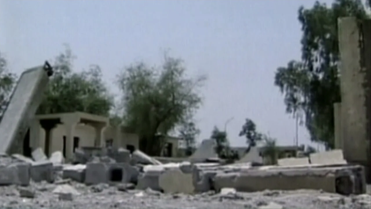 Ghosts of Abu Ghraib (2007) movie still: establishing — A pile of grey concrete rubble sits in the foreground of a leveled lot. Several damaged…