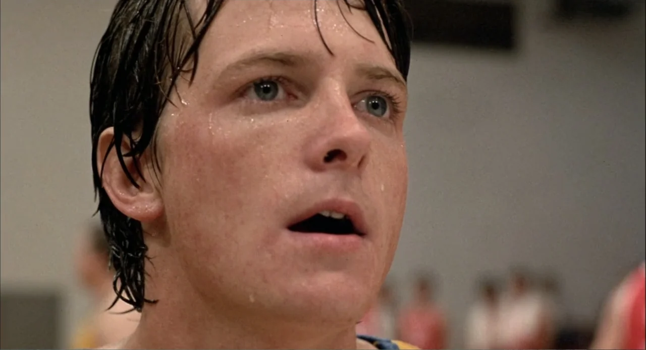 Teen Wolf (1985) movie still: closeup — An indoor gymnasium with out-of-focus figures present in the distant background. A young man with damp,…