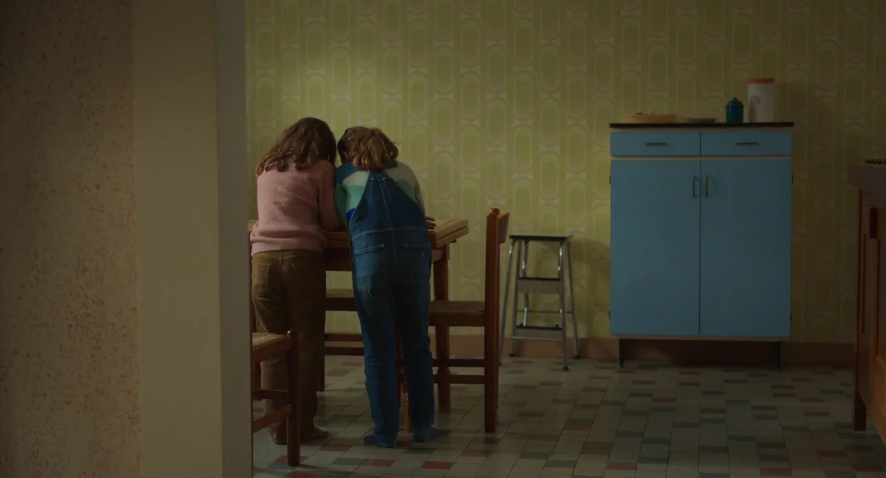 Petite Maman (2021) movie still: fullbody — A 1970s interior features olive green geometric wallpaper and vintage blue cabinets. Two young children…