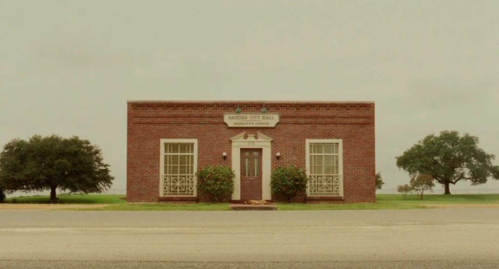 The Ladykillers (2004) movie still: establishing — A red brick, single-story government building stands alone in a flat, grassy landscape. Flanking…
