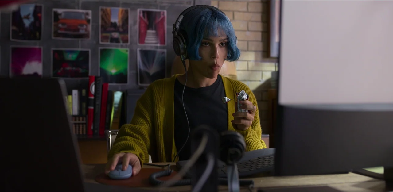 Kimi (2022) movie still: medium — A person sits at a desk with computer peripherals and photographic prints on the wall. The subject wears…