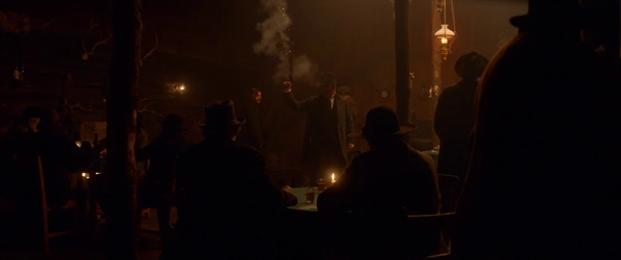 Never Grow Old (2019) movie still: wide — A dimly lit, wood-walled building interior features several figures seated at tables. One man stands and…