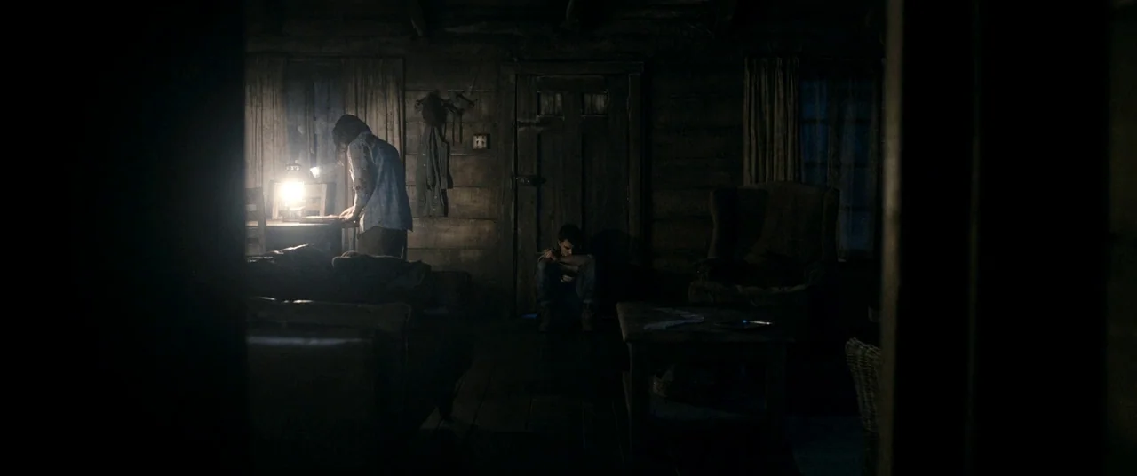 Evil Dead (2013) movie still: wide — A dimly lit cabin interior with wooden walls and rustic furniture. Two individuals are situated in different…