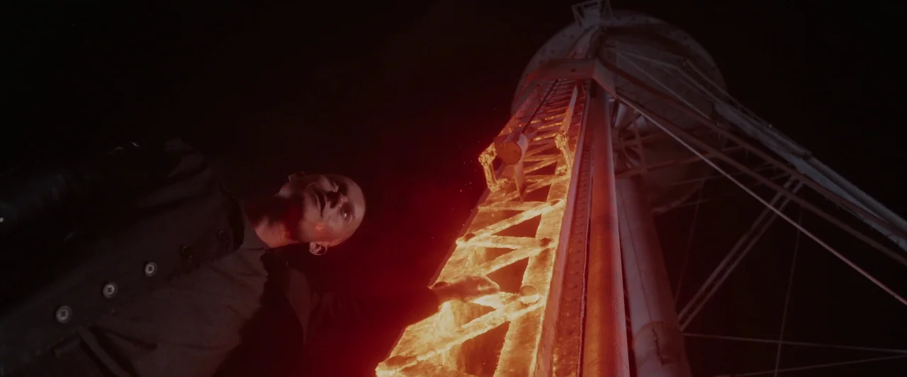 Iron Man 3 (2013) movie still: medium — A dark night scene features a metal water tower frame illuminated by intense firelight. A man stands at the…