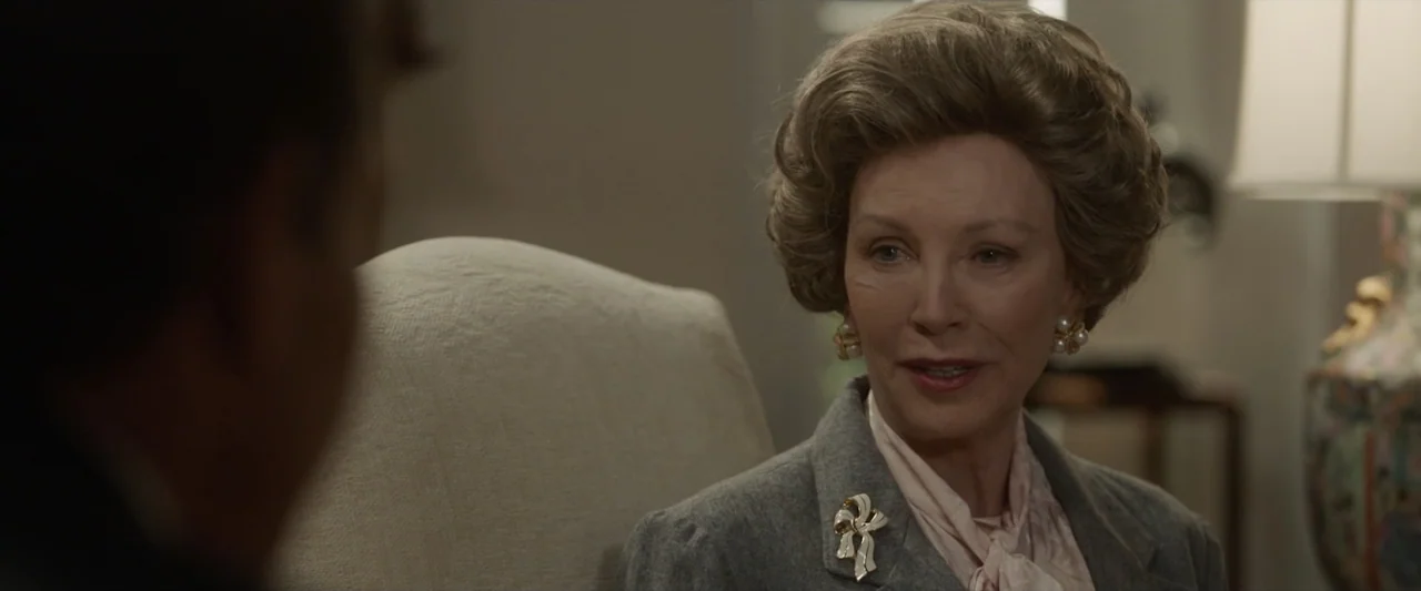Reagan (2024) movie still: medium — A well-lit interior room contains a woman in formal business attire. She sits in a chair and speaks to an…