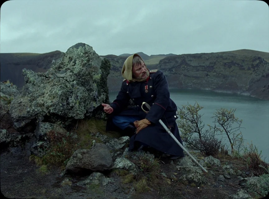Jauja (2014) movie still: medium — A man in a military coat sits on a mossy volcanic rock overlooking a lake. The man crouches on the uneven ground…