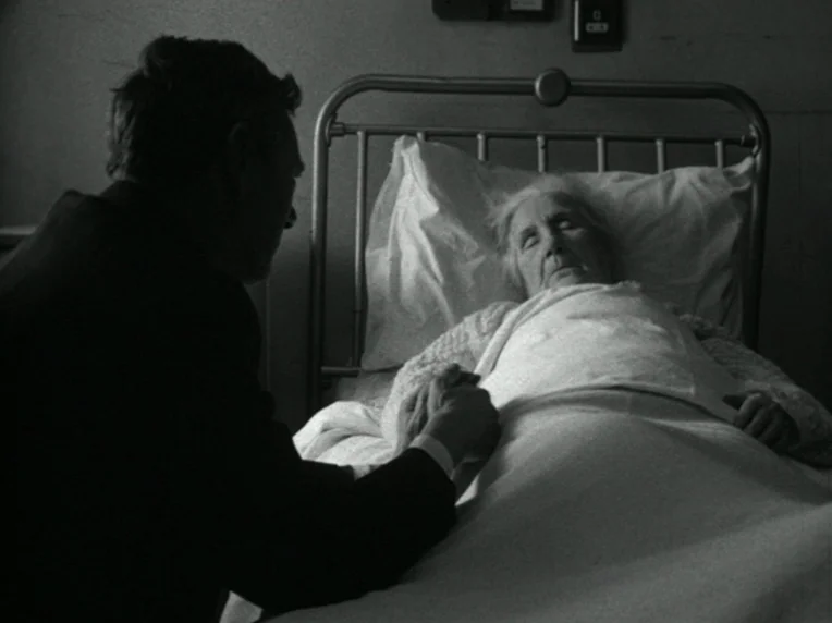 Death and Transfiguration (1983) movie still: medium — A dark hospital room contains a metal-framed bed with white bedding and pillows. An elderly…