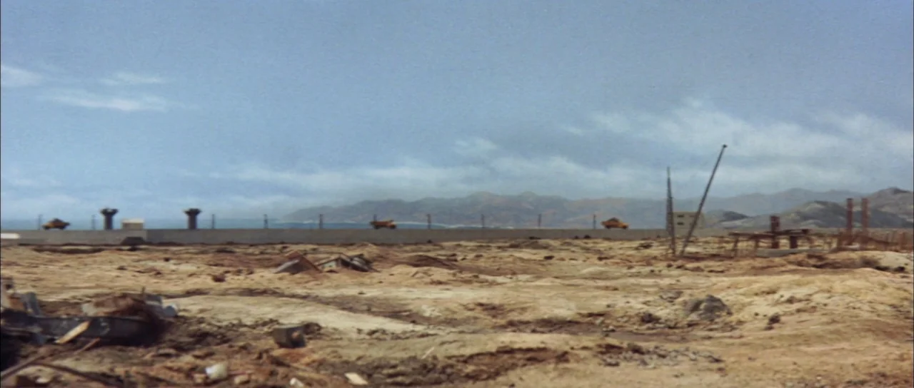 Mothra Vs Godzilla (1964) movie still: establishing — A vast, barren construction site features uneven, dirt-covered ground under a clear, pale sky.…