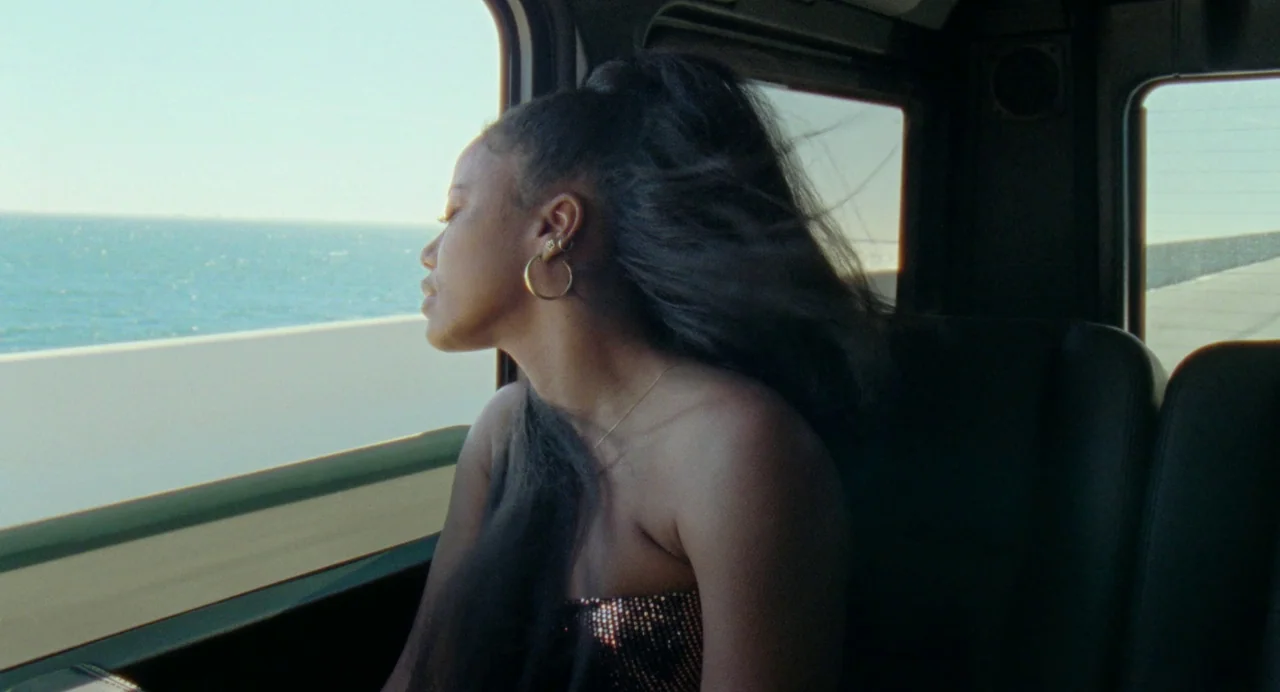 Zola (2020) movie still: medium — The interior of a vehicle moving along a coastal road next to the sea. A young woman looks out of the window as…