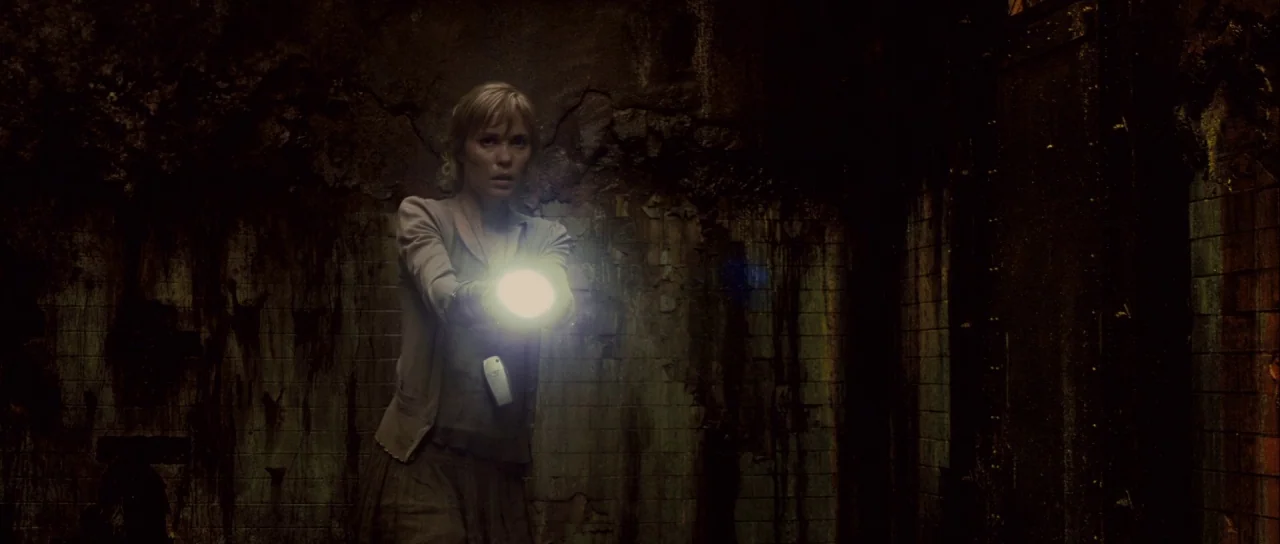 Silent Hill movie still: medium — A dark subterranean space features grimy, wet tiled walls covered in deep mineral staining. A woman standing in…