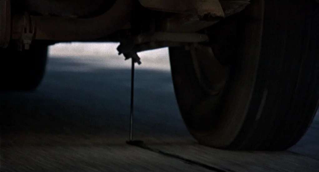 Family Plot (1976) movie still: closeup — The underside of a vehicle frame shows a metal rod dragging against asphalt. The tire is visible in the…