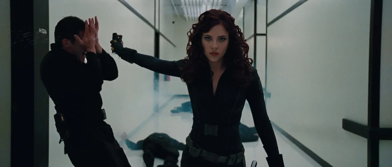Iron Man 2 (2010) movie still: medium — A sterile, white-walled corridor with black trim and fallen bodies on the floor. A woman in a black tactical…