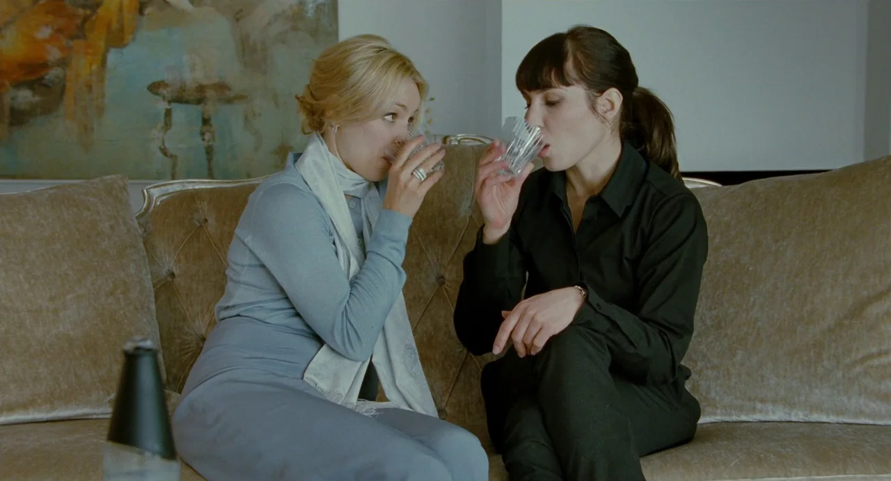 Passion (2012) movie still: medium — A refined interior space features a tan velvet sofa and an abstract painting. Two women in contrasting outfits…