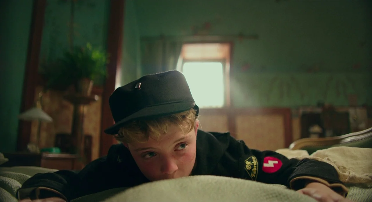 Jojo Rabbit (2019) movie still: medium — A cramped attic room features vintage patterned wallpaper and a lone potted plant. A young boy lies on his…